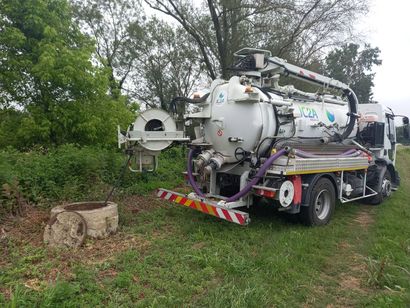 Un gros camion aspirateur blanc est stationné sur le bord d'une route herbeuse, à côté d'arbres et de buissons.