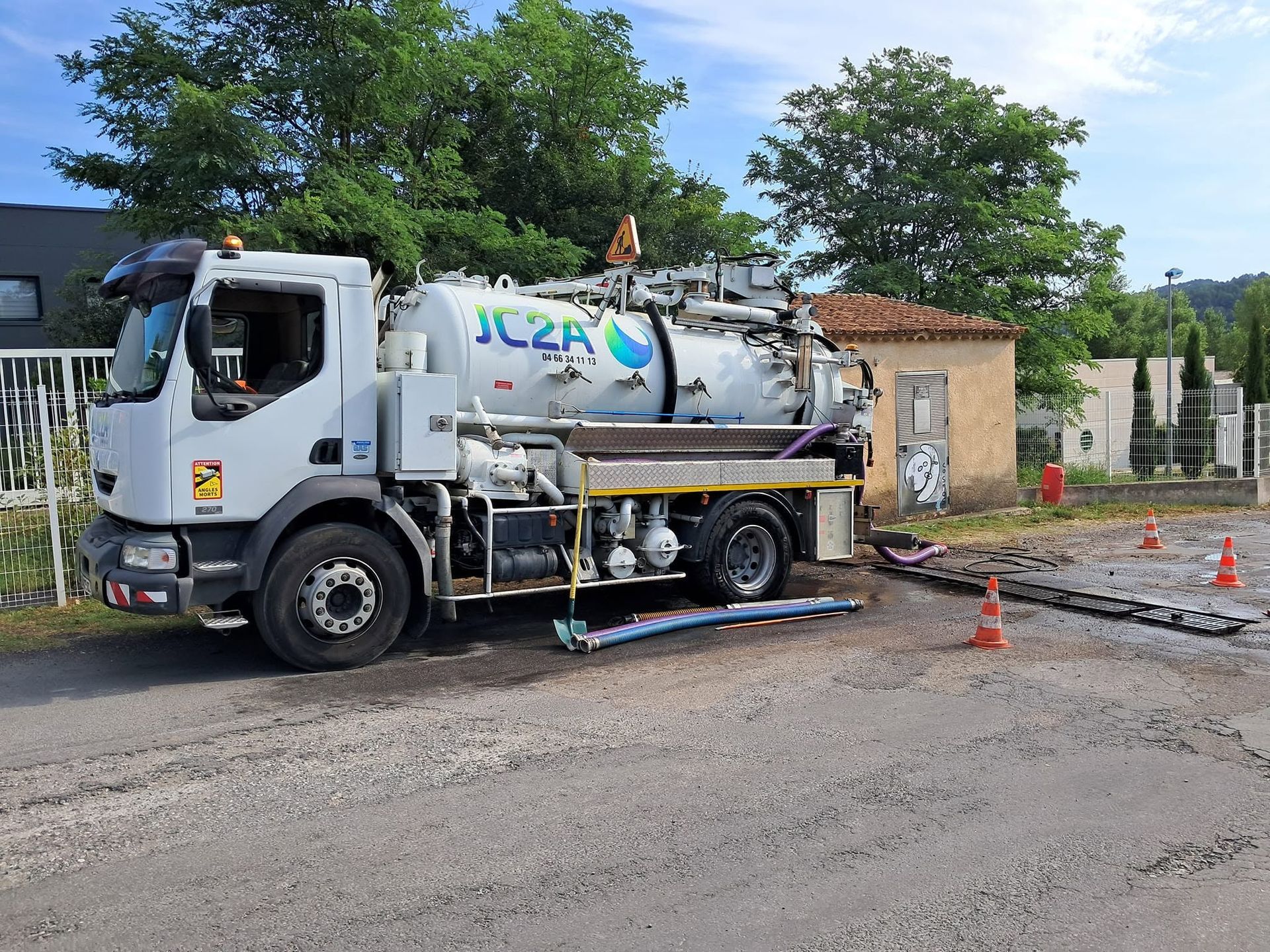 Camion aspirateur blanc sur une route longeant un chantier, avec des cônes orange et une chaussée mouillée.