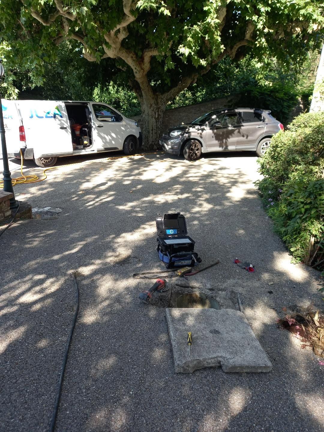 Allée avec une camionnette blanche et un SUV gris garés sous un grand arbre, avec une petite malette d'équipement au premier plan.