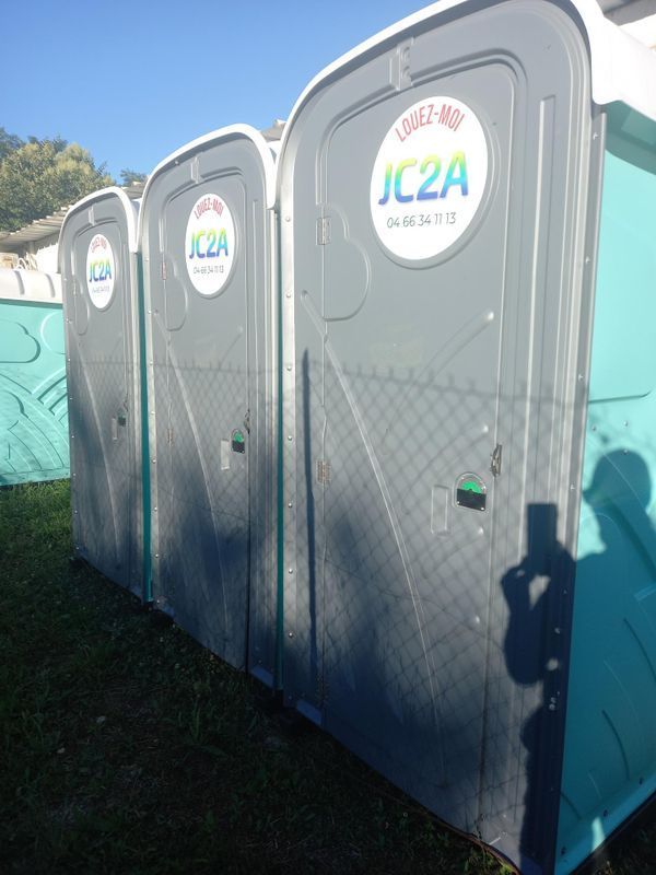 Rangée de toilettes portables à l'extérieur, avec des portes grises et des panneaux latéraux bleus, sous un soleil éclatant.