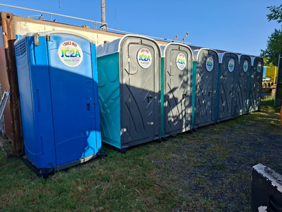 Une rangée de toilettes portables bleues et grises, installées à l'extérieur sur l'herbe sous un ciel dégagé.
