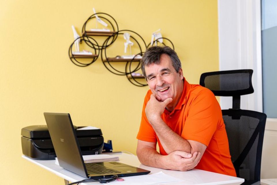 Ein Mann in einem orangefarbenen Hemd lächelt an einem Schreibtisch mit Laptop, Drucker und gelber Wand.