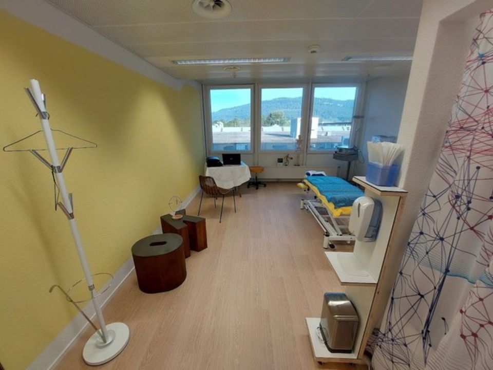 Ein lichtdurchfluteter Untersuchungsraum mit Untersuchungsliege, Garderobe und Fensterblick auf die Berge.