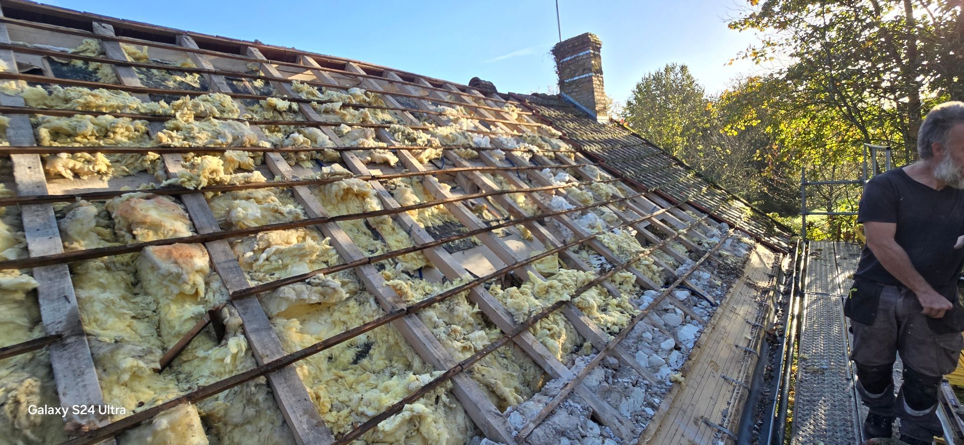 Un homme se tient debout sur un échafaudage à côté d'un toit en construction, laissant apparaître des poutres en bois et une isolation jaune.