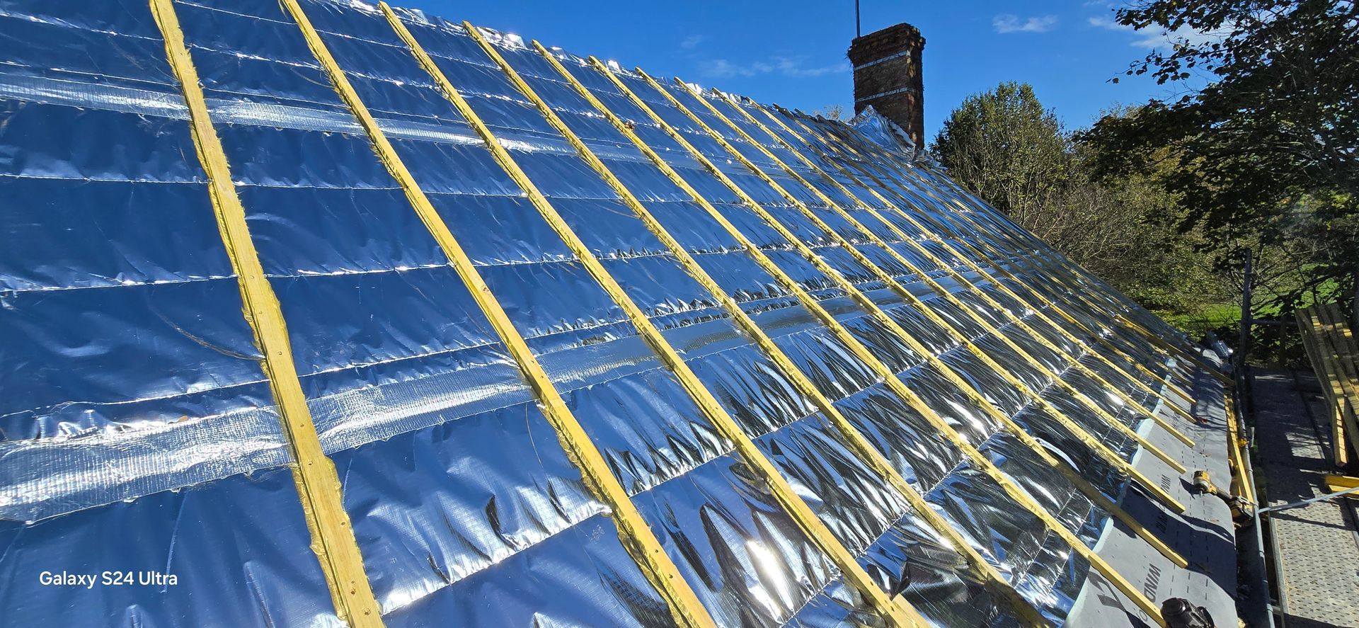 Un toit en pente en construction, avec des chevrons verticaux en bois installés sur une couche d'isolant argenté réfléchissant.