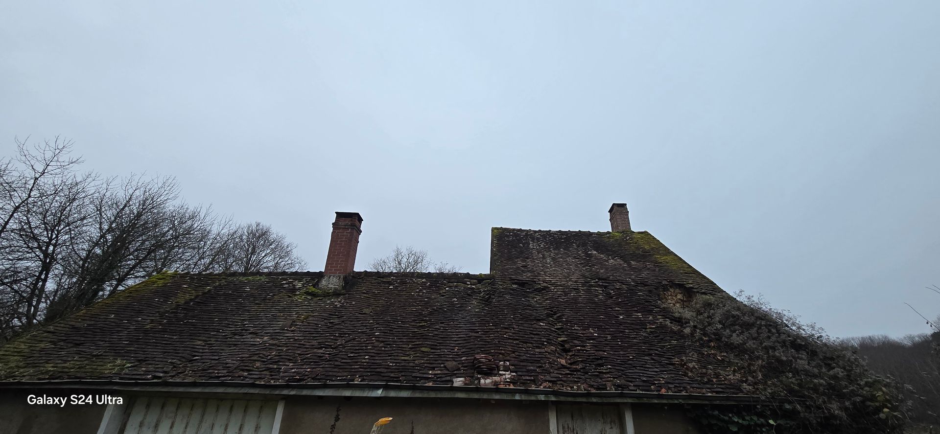 Un vieux toit de tuiles moussus avec deux cheminées.