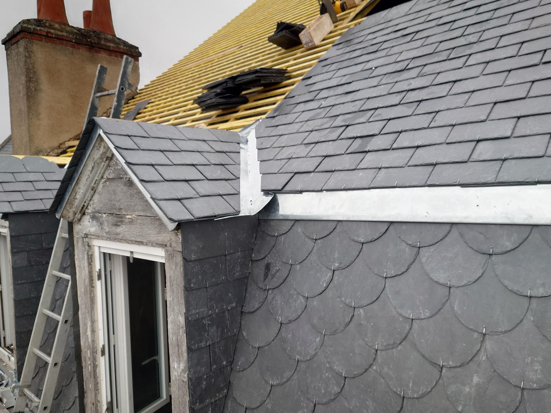 Toiture d'un bâtiment en cours de réparation, laissant apparaître les tuiles d'ardoise et les solins métalliques.