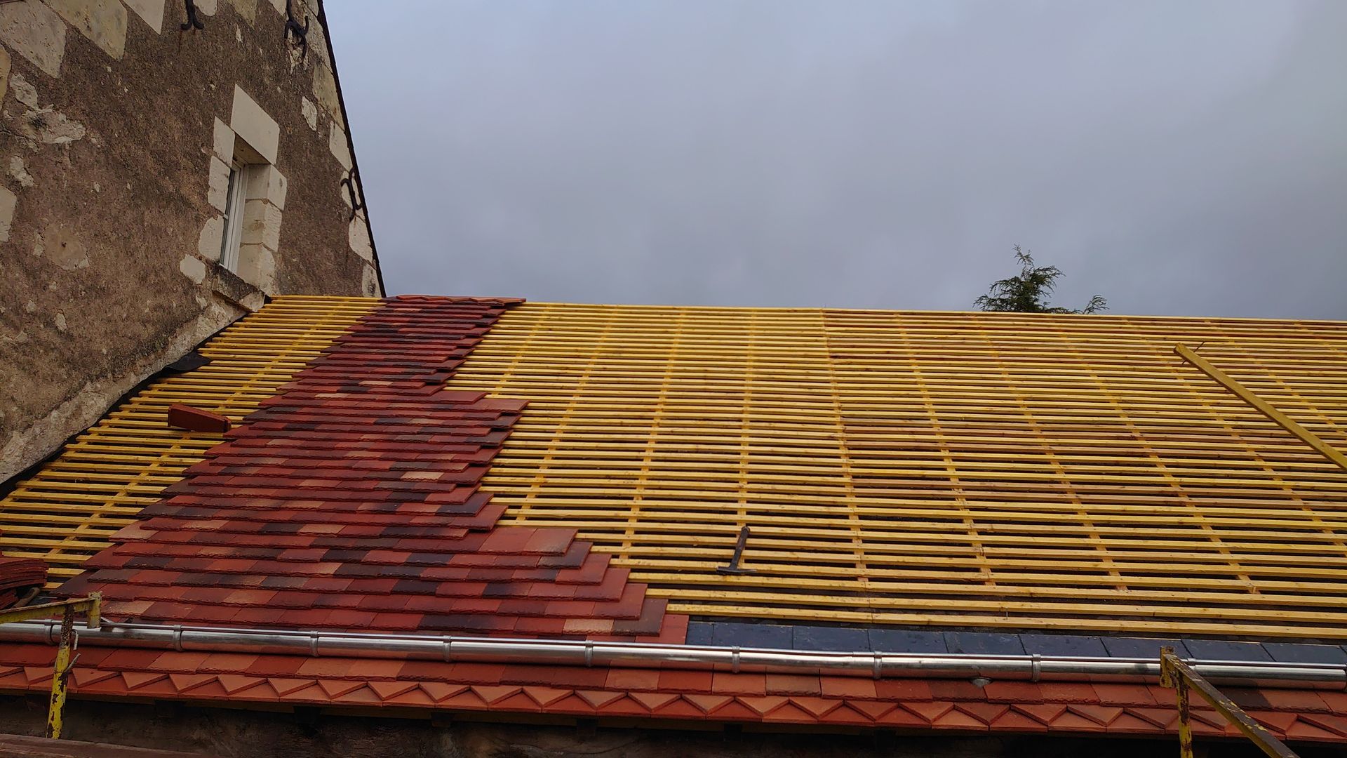 Un toit partiellement couvert de tuiles, en construction, avec des liteaux en bois et des tuiles en terre cuite rouge, adossé à un vieux mur de pierre.