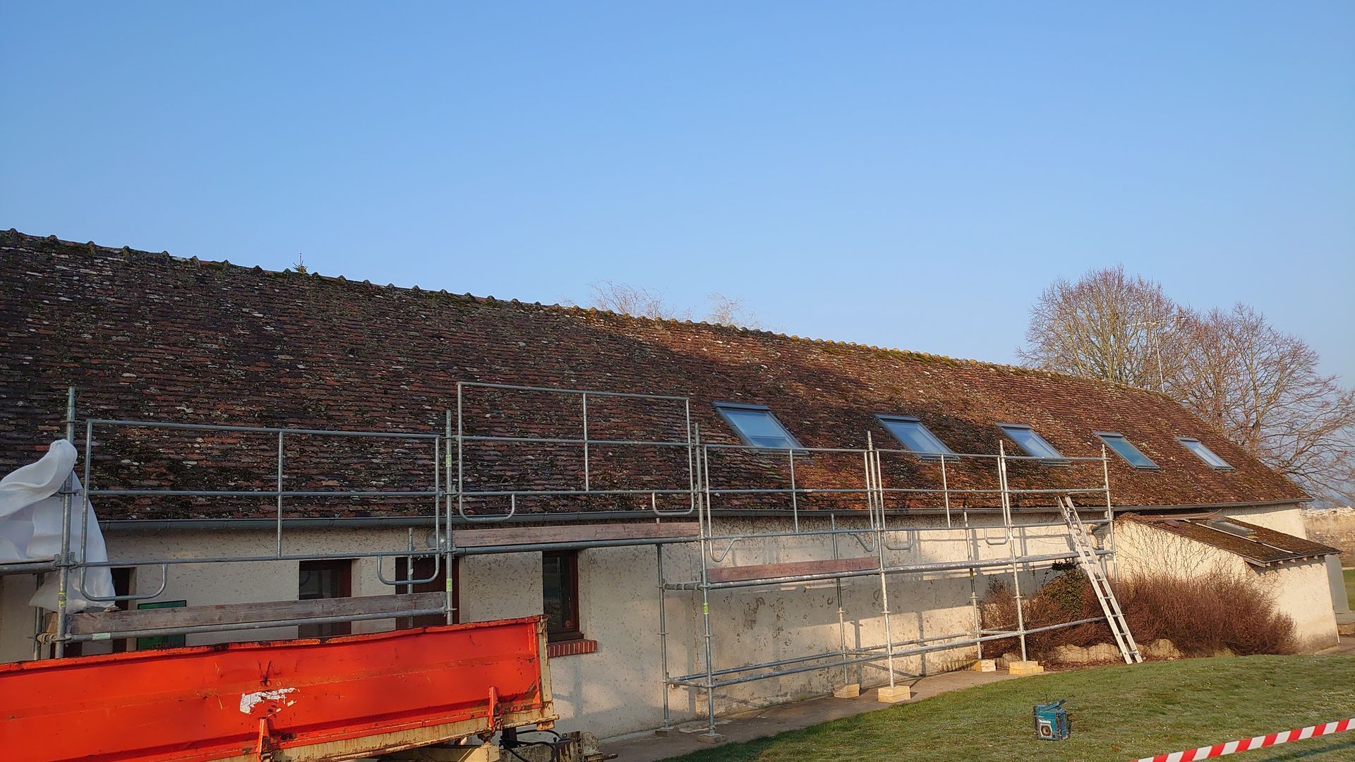 Un long bâtiment au toit de tuiles moussu, en cours de rénovation, entouré d'échafaudages métalliques.