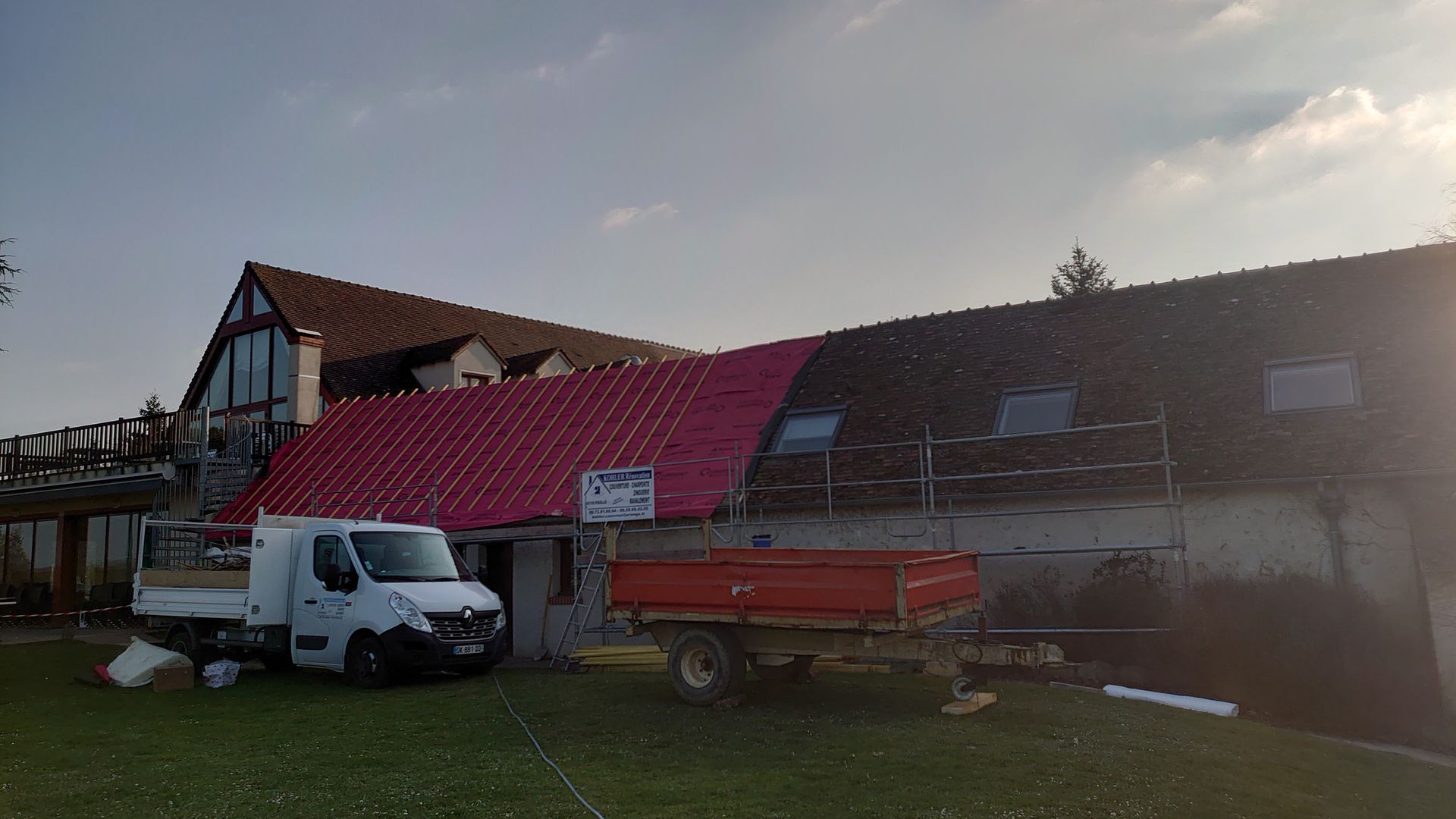 Travaux de construction sur le toit d'une maison présentant une sous-couche rose vif partiellement installée, avec une camionnette blanche et une remorque.