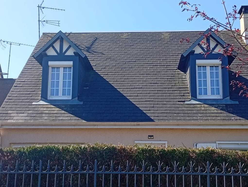 Deux lucarnes sur un toit en bardeaux foncés ; une maison beige avec une haie verte et une clôture en fer forgé noir.