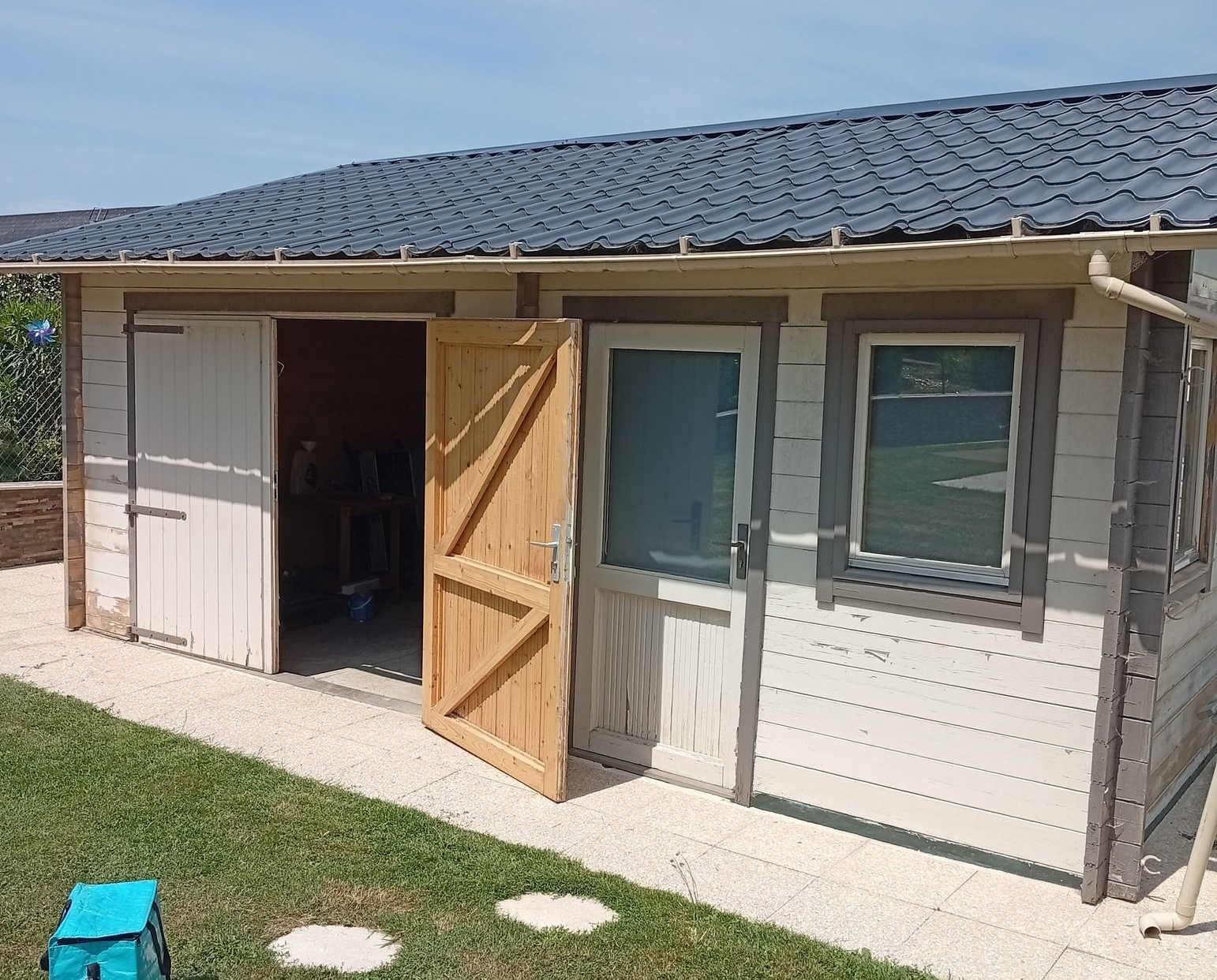 Abri en bois avec portes ouvertes sur une zone herbeuse, conteneur bleu clair au premier plan.
