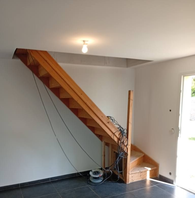 Escalier en bois menant à une ouverture dans le plafond, fils suspendus, dans un espace intérieur inachevé.