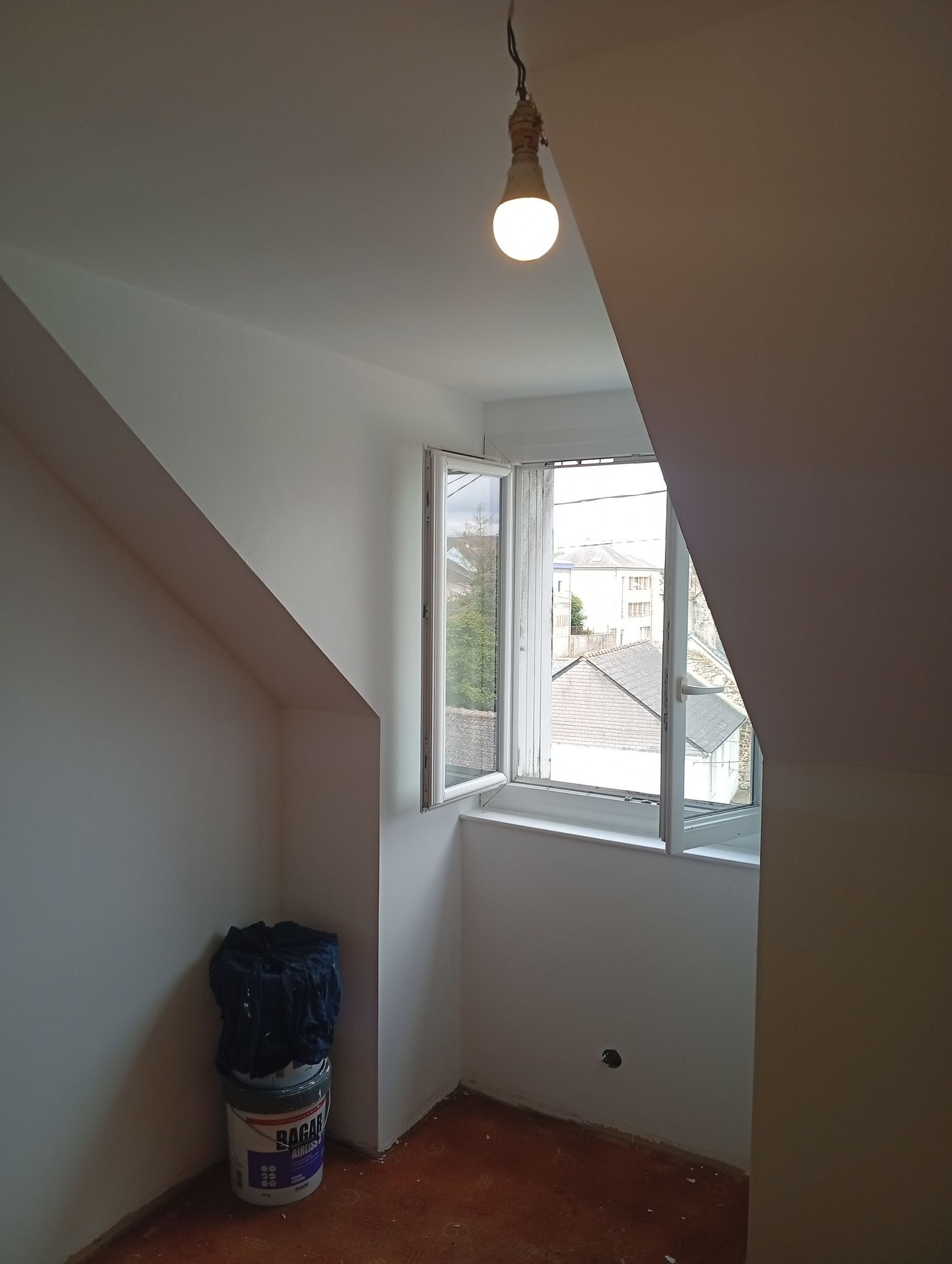 Petite pièce avec des murs inclinés, une fenêtre et une ampoule suspendue ; seau de peinture et sac-poubelle dans le coin.