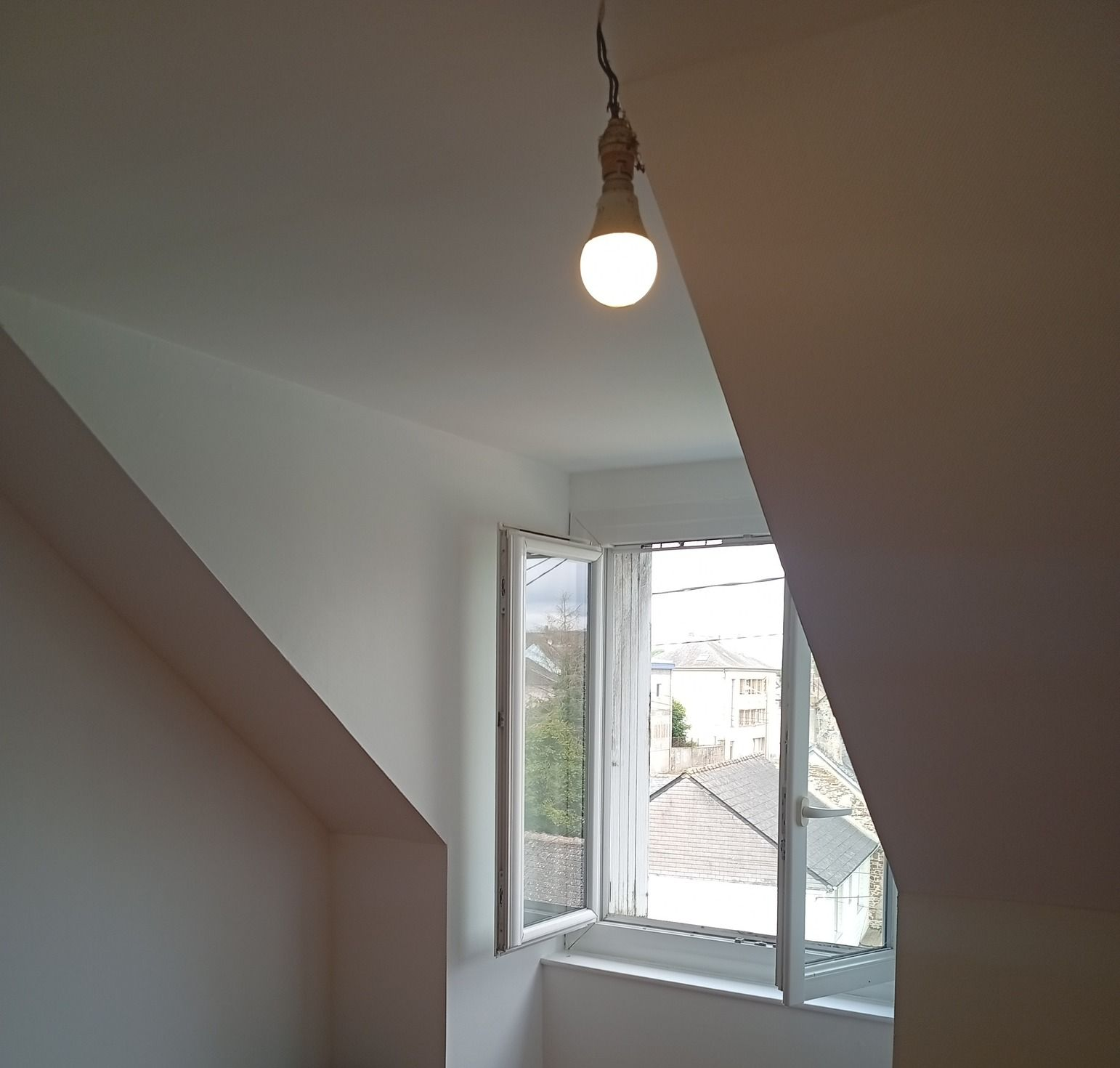 Une ampoule allumée est suspendue dans une pièce avec fenêtre. Murs blancs et plafond incliné.