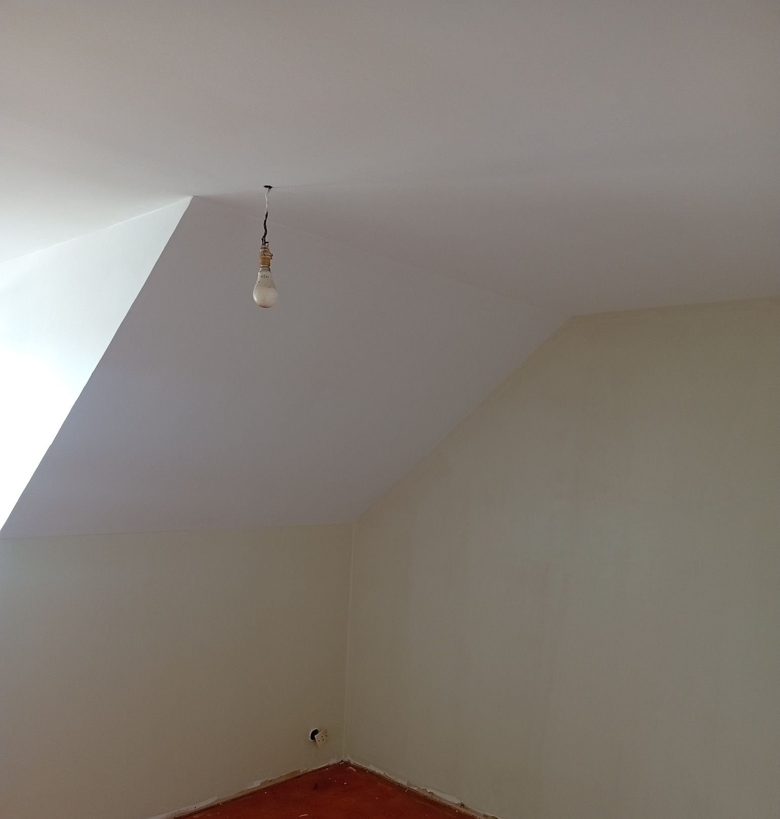 Chambre blanche avec plafond en pente sur lequel une ampoule nue est suspendue et plancher en bois brut.