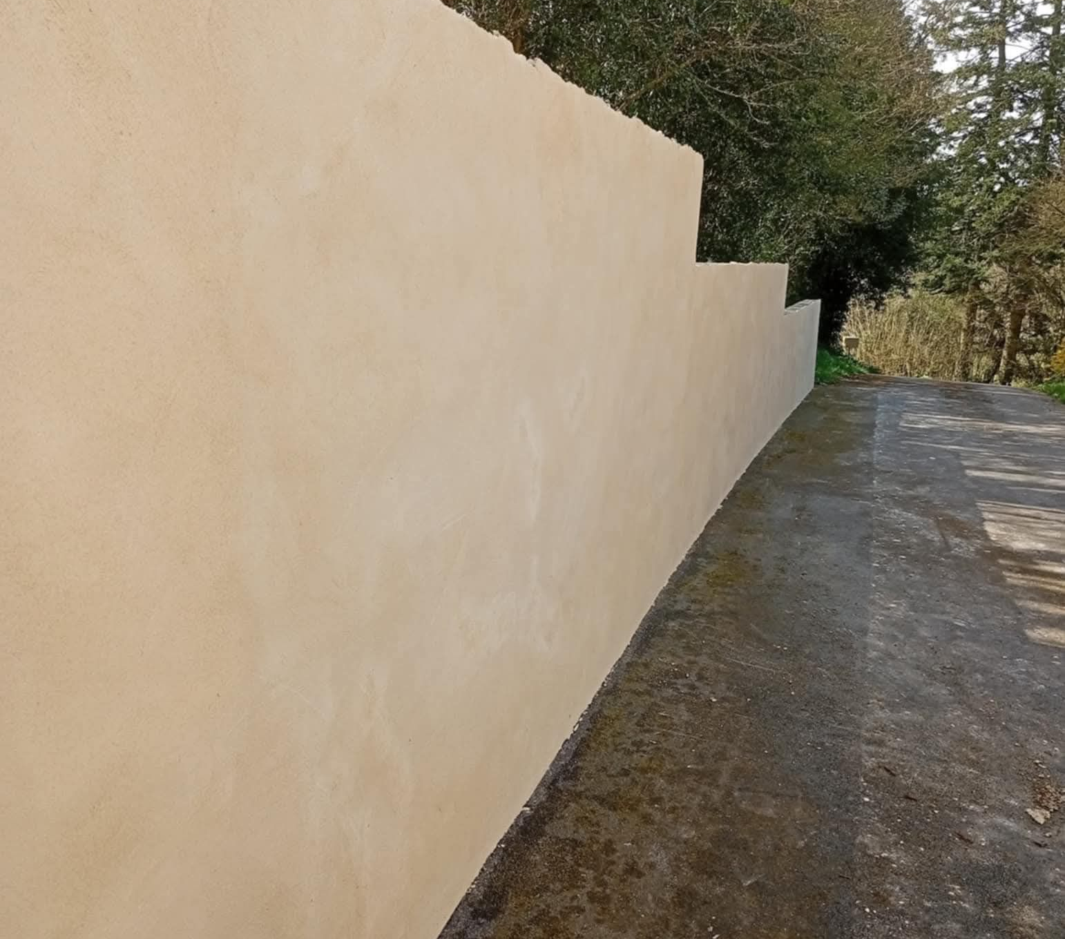 Mur en stuc beige le long d'une allée gris foncé, arbres en arrière-plan.