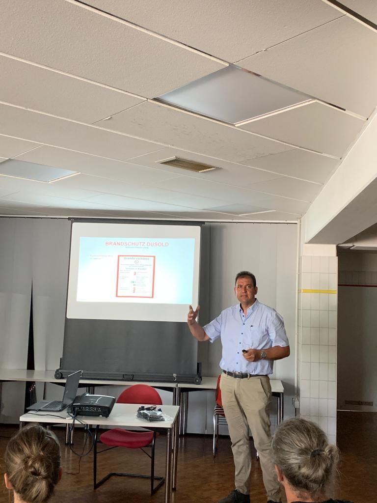 Ein Mann hält in einem hell erleuchteten Raum eine Präsentation und zeigt dabei auf ein projiziertes Diagramm.