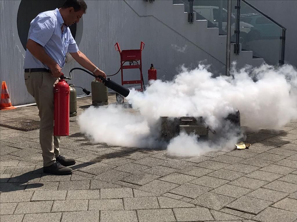 Eine Person löscht mit einem Feuerlöscher einen Brand in einer Kiste, wodurch draußen eine große Rauchwolke entsteht.