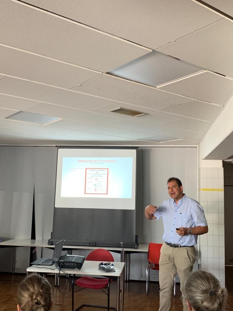 Ein Mann hält eine Präsentation und zeigt auf ein projiziertes Diagramm. In einem Klassenzimmer.