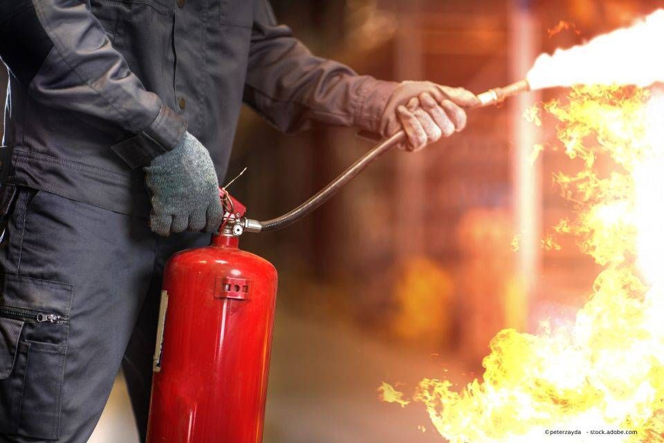 Eine Person in blauem Overall und Handschuhen benutzt einen Feuerlöscher, um ein Feuer zu bekämpfen.