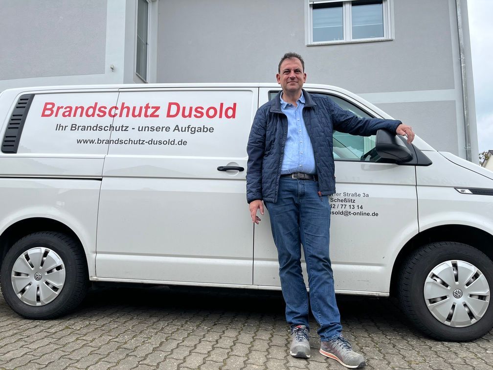 Ein Mann steht neben einem weißen Lieferwagen mit der Aufschrift „Brandschutz Dusold“. Im Hintergrund ist ein Gebäude zu sehen.