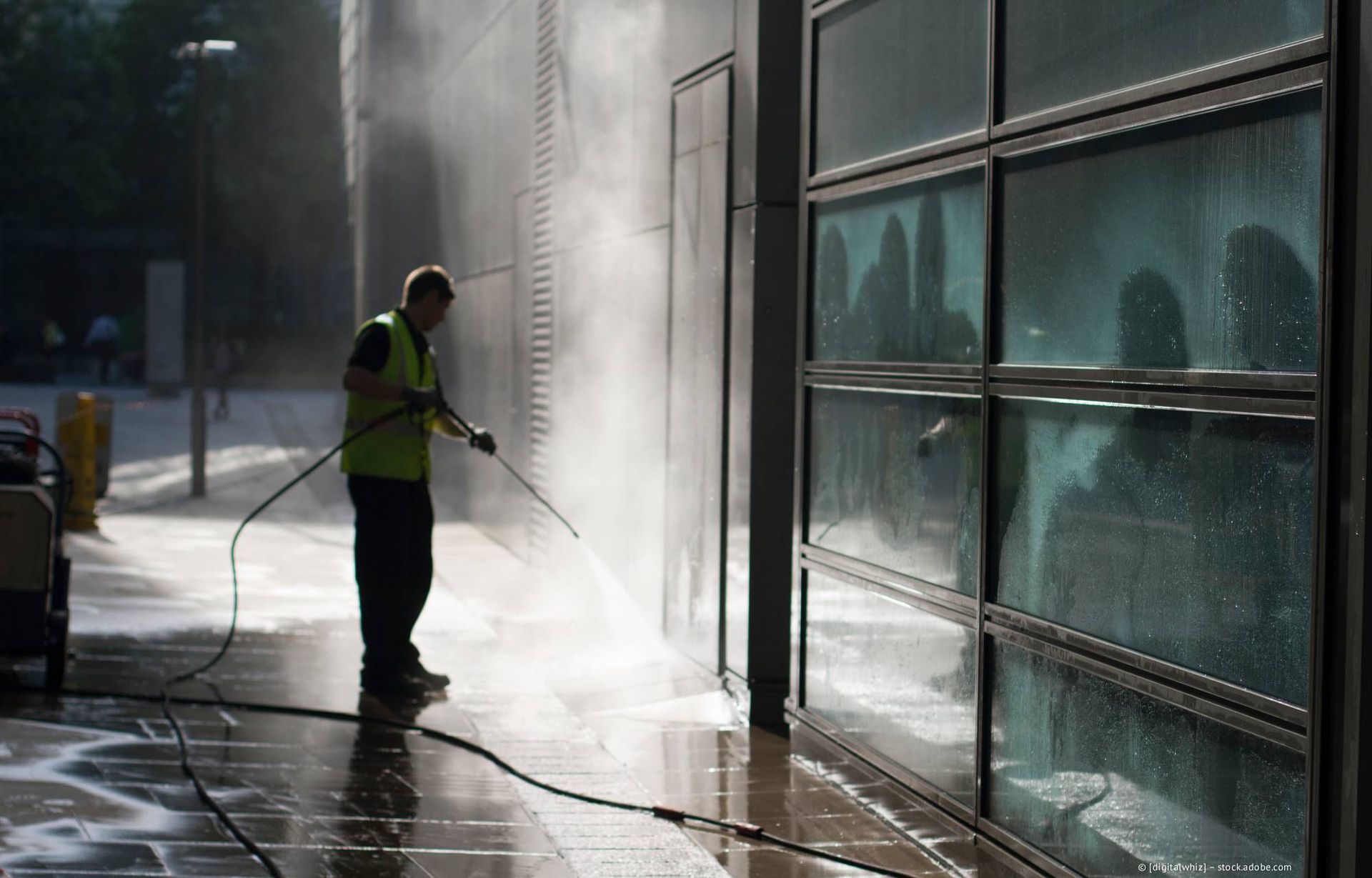 Ein Mann in einer grünen Weste reinigt mit einem Hochdruckreiniger die Fassade eines Gebäudes mit Glasfenstern und Betonboden.