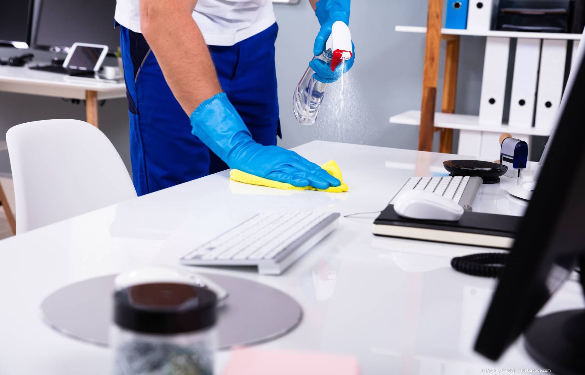 Eine Person mit blauen Handschuhen desinfiziert einen weißen Büroschreibtisch mit einer Sprühflasche und einem gelben Tuch.