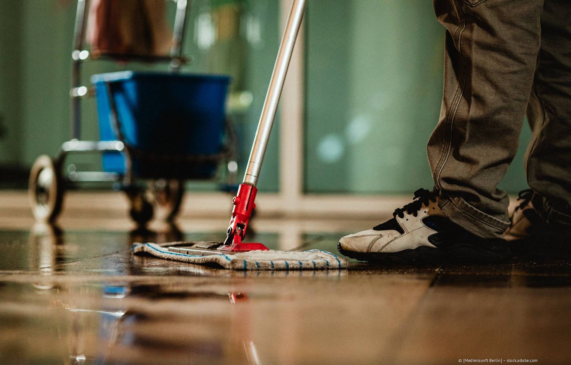 Eine Person wischt einen nassen, spiegelnden Boden; im Hintergrund ist ein Reinigungswagen zu sehen.