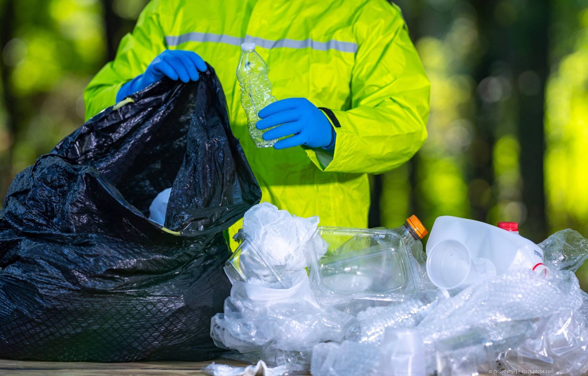 Person in grünem Regenmantel und blauen Handschuhen steckt Plastikflasche in schwarzen Müllsack, im Freien.