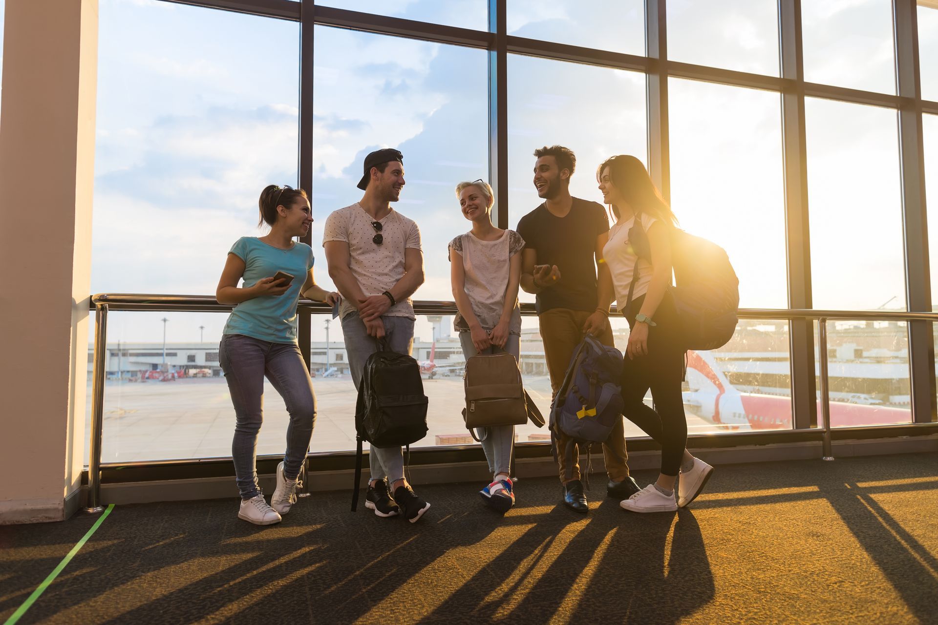 Groupe de voyageurs discutant devant la baie vitrée d'un aéroport, au soleil couchant