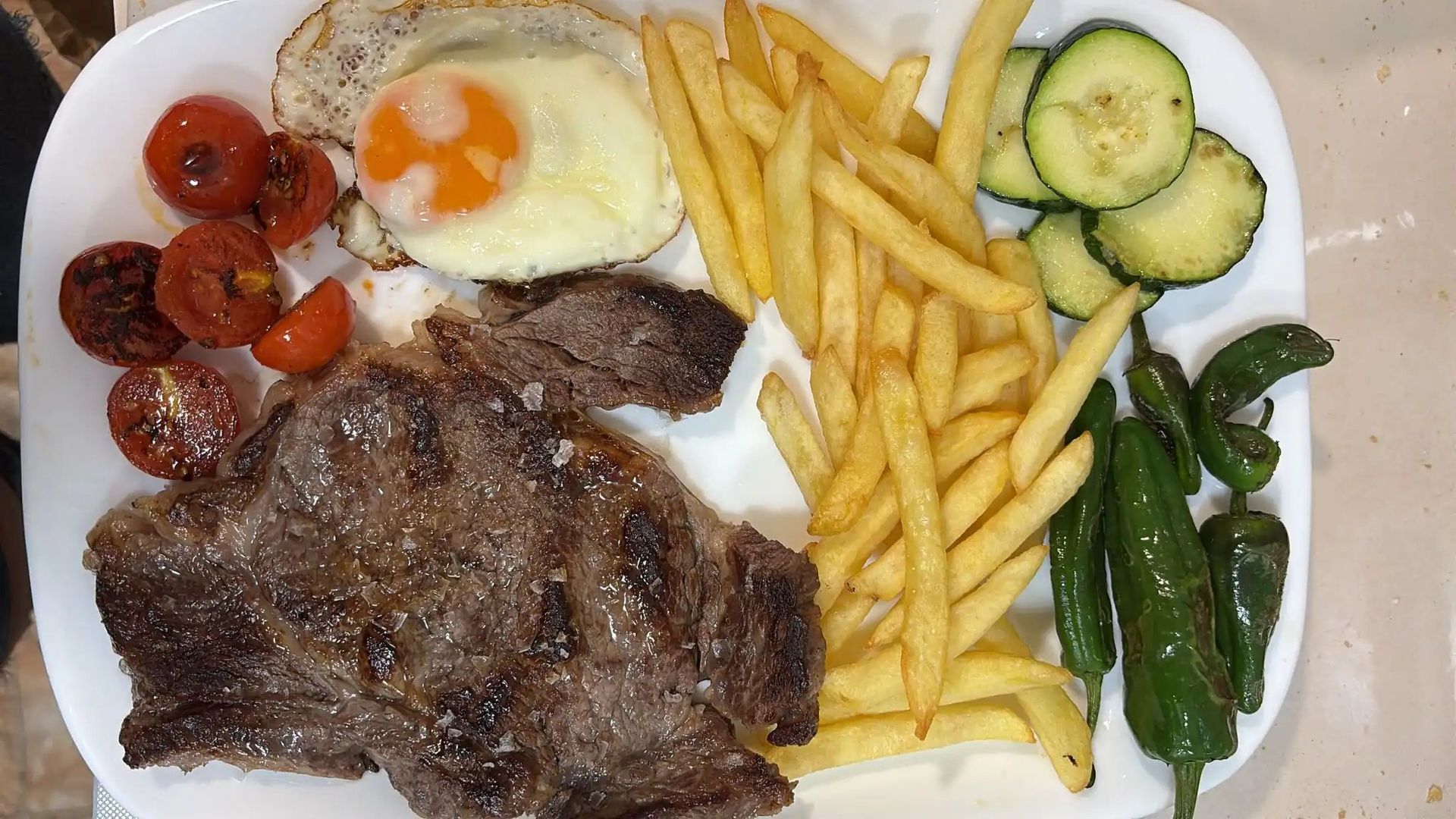Plato de comida: filete, huevo frito, patatas fritas, tomates, calabacín, pimientos.