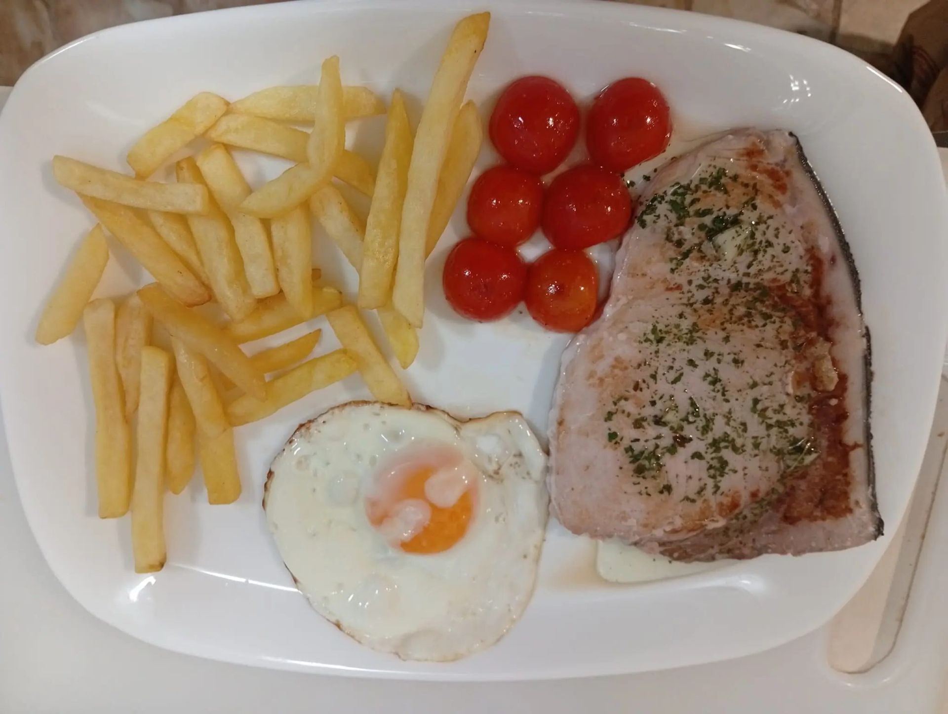 Plato con patatas fritas, tomates cherry, huevo frito y un trozo de pescado a la plancha.