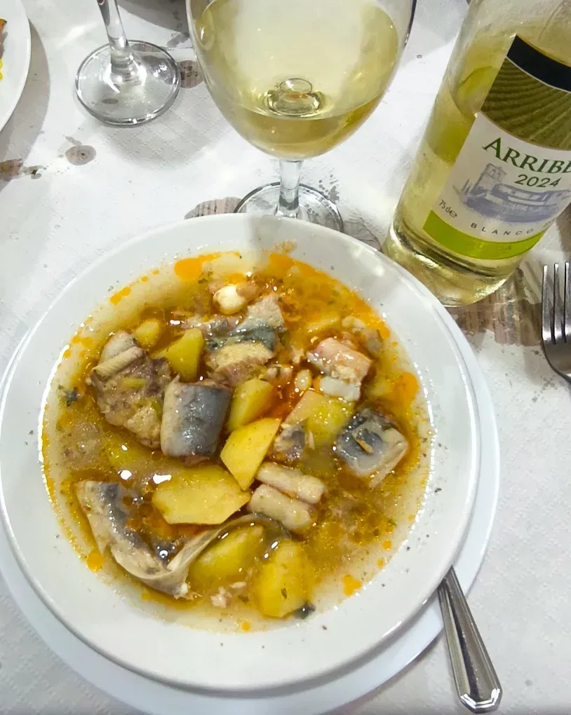 Cuenco de guiso de pescado con patatas, judías, botella de vino y copa sobre mantel blanco.