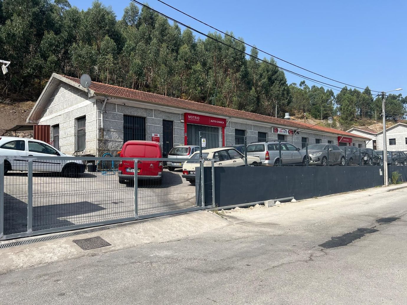 Uma fileira de prédios comerciais com carros estacionados atrás de uma cerca de metal, árvores ao fundo, dia ensolarado.