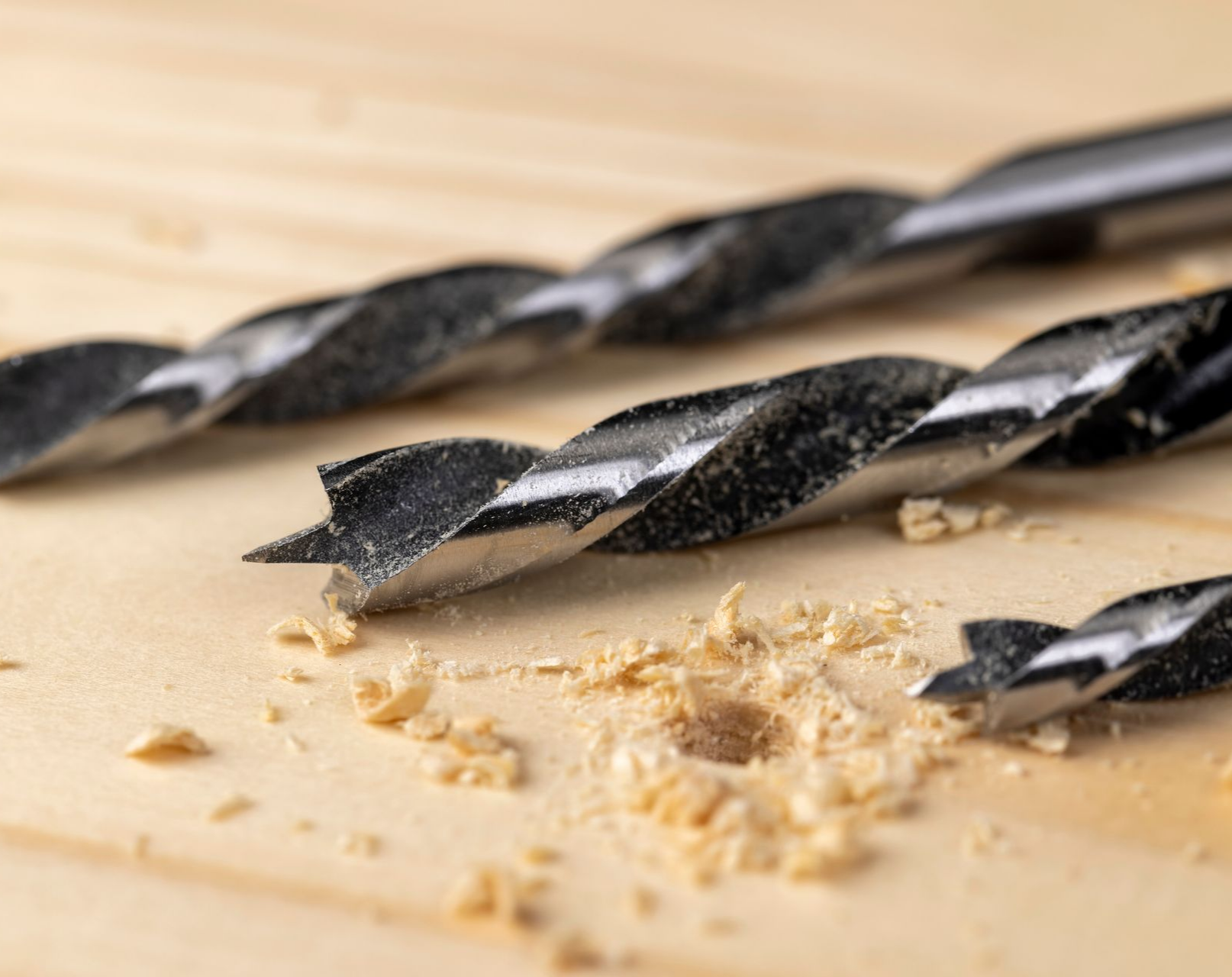 Trois mèches de foret noires sur une surface en bois clair, l'une d'elles devant un trou avec des copeaux de bois.