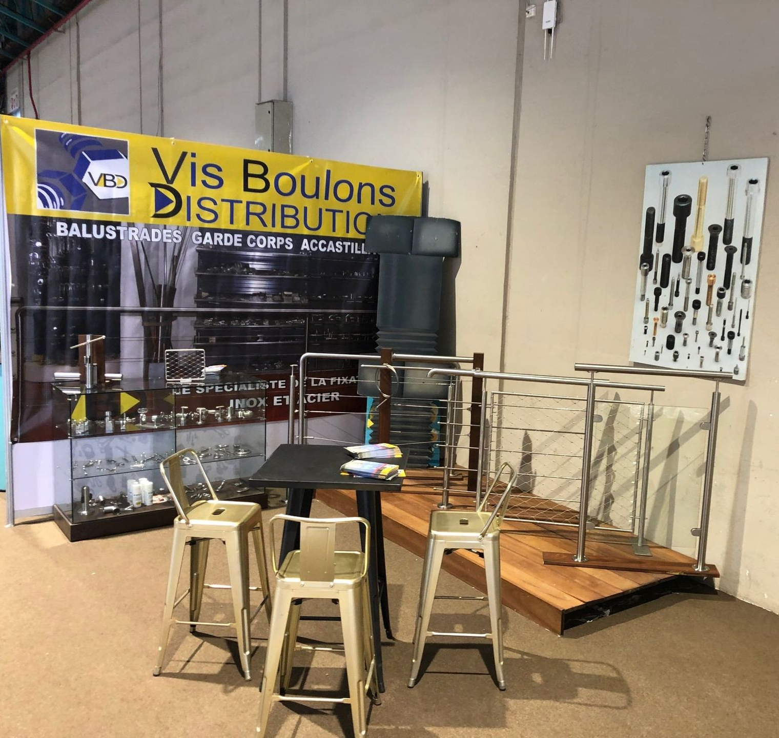 Un stand d'exposition présente des garde-corps en acier inoxydable, des tabourets de bar et des échantillons de produits portant le logo VIS BOULONS DISTRIBUTION.