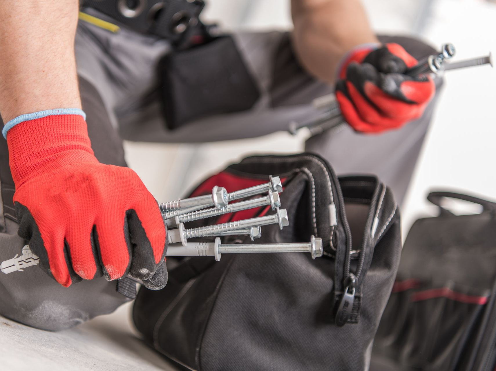 Personne portant des gants rouges tenant des boulons, agenouillée près d'une sacoche à outils.