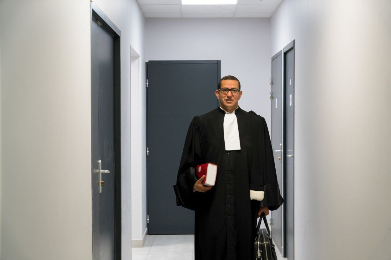 Maître Zouaoui dans un couloir, en robe d'avocat, avec un livre dans la main droite