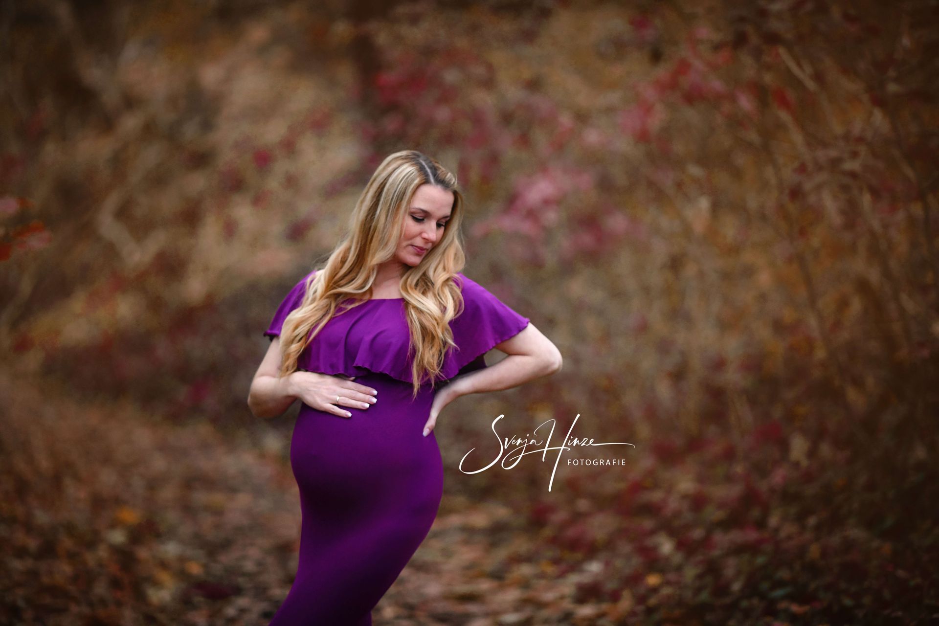 Eine schwangere Frau in einem lila Kleid steht im Wald und hält ihren Bauch.