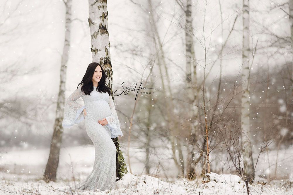 Eine schwangere Frau in einem weißen Kleid steht im Schnee neben einem Baum.