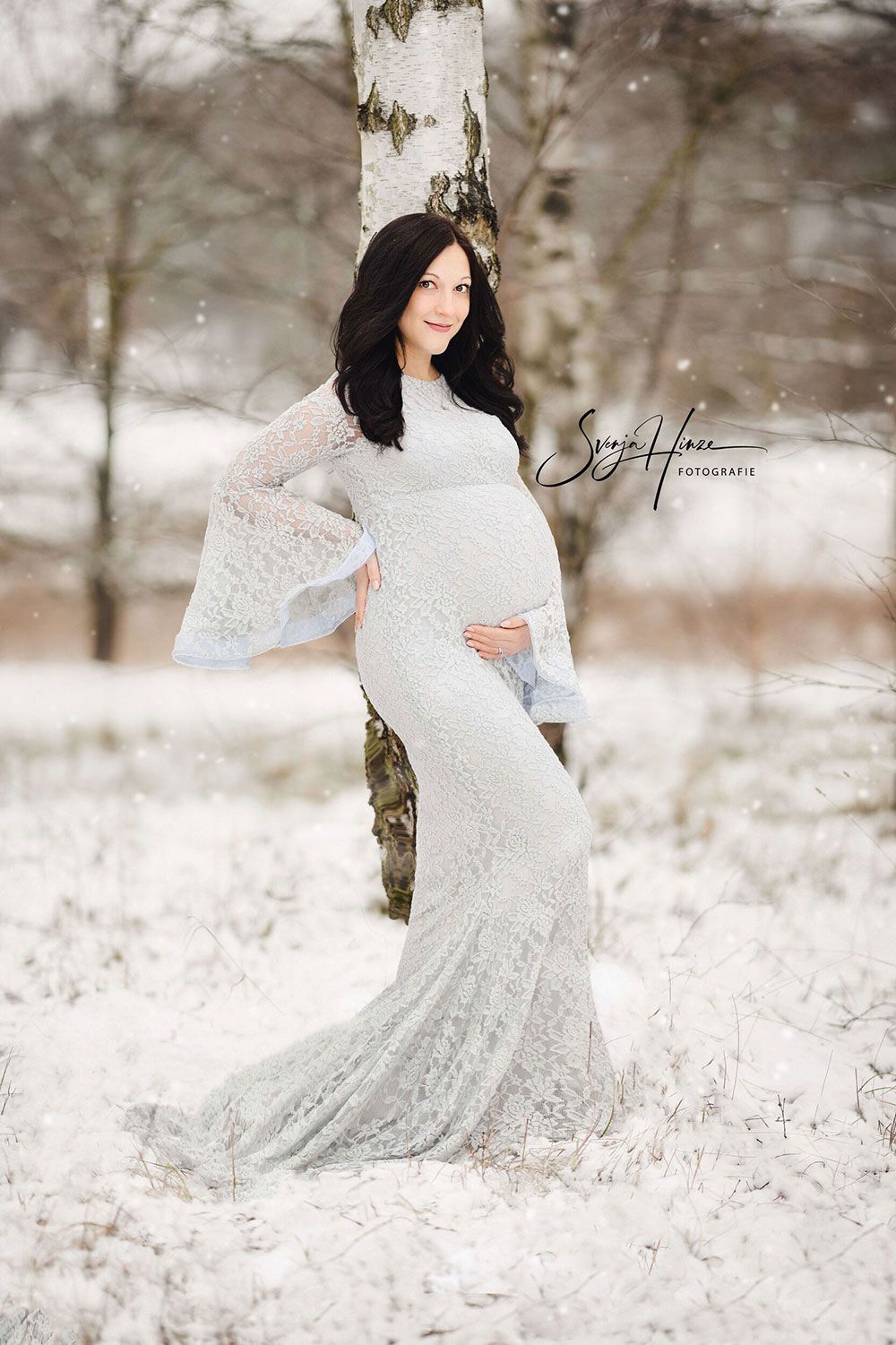 Eine schwangere Frau in einem weißen Kleid steht im Schnee neben einem Baum.