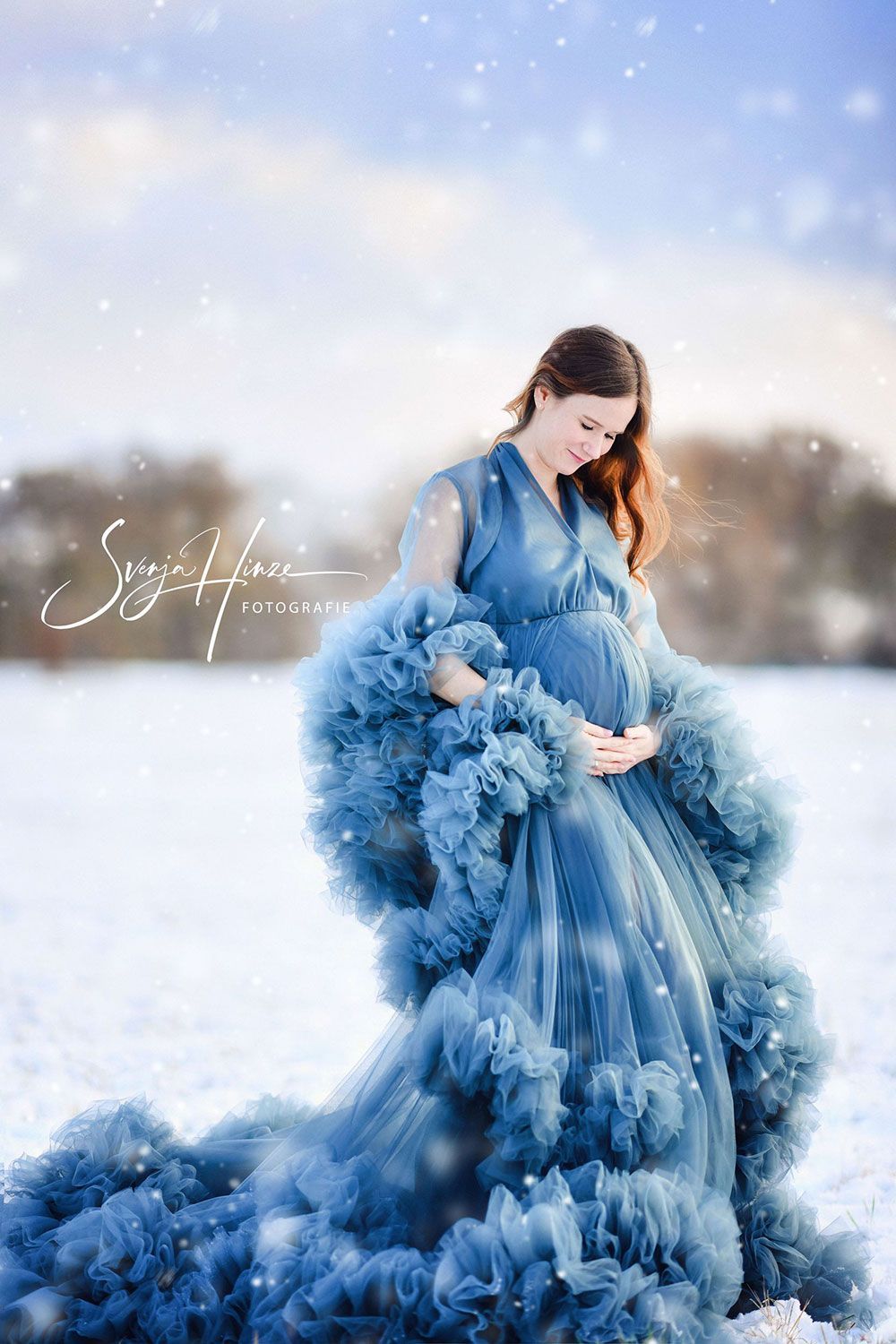 Eine schwangere Frau in einem blauen Kleid steht im Schnee.