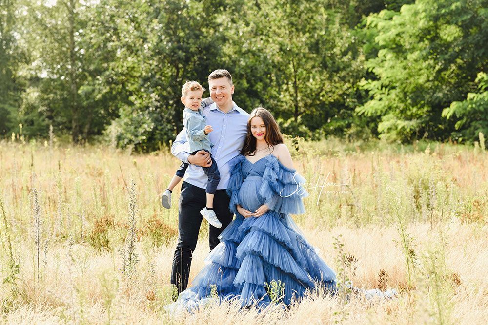 Eine schwangere Frau in einem blauen Kleid steht mit ihrem Mann und ihrem Sohn auf einem Feld.