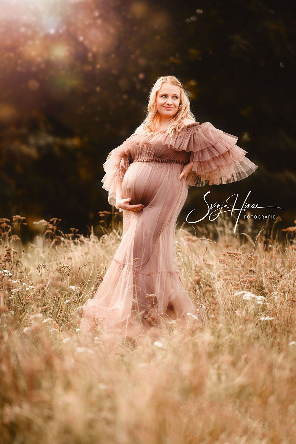 Eine schwangere Frau in einem langen Kleid steht auf einem Feld und hält ihren Bauch.