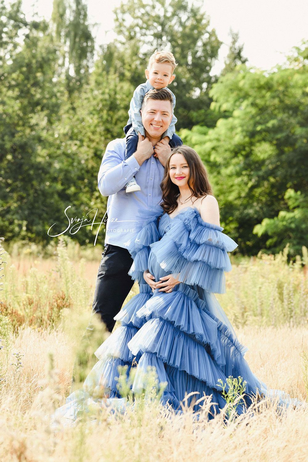 Eine schwangere Frau in einem blauen Kleid steht mit ihrem Mann und ihrem Sohn auf einem Feld.