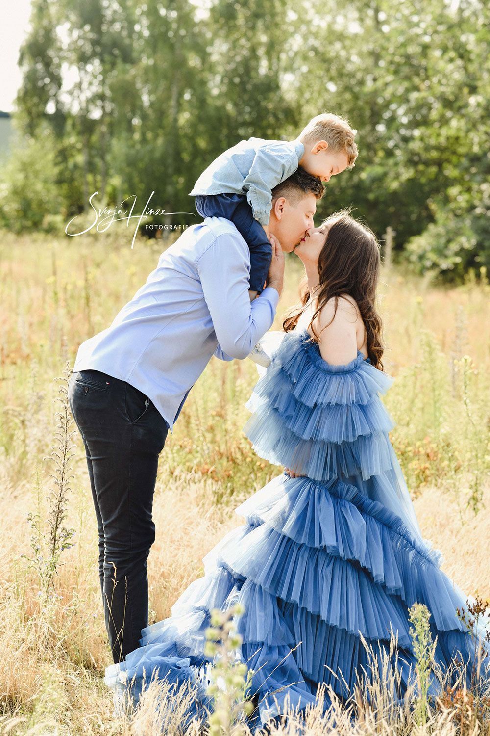 Ein Mann küsst eine Frau auf die Wange, während er auf einem Feld ein Kind auf seinen Schultern trägt.