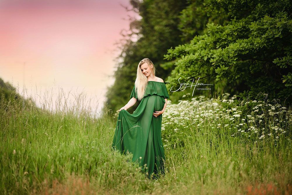 Eine schwangere Frau in einem grünen Kleid steht auf einem Feld mit hohem Gras.