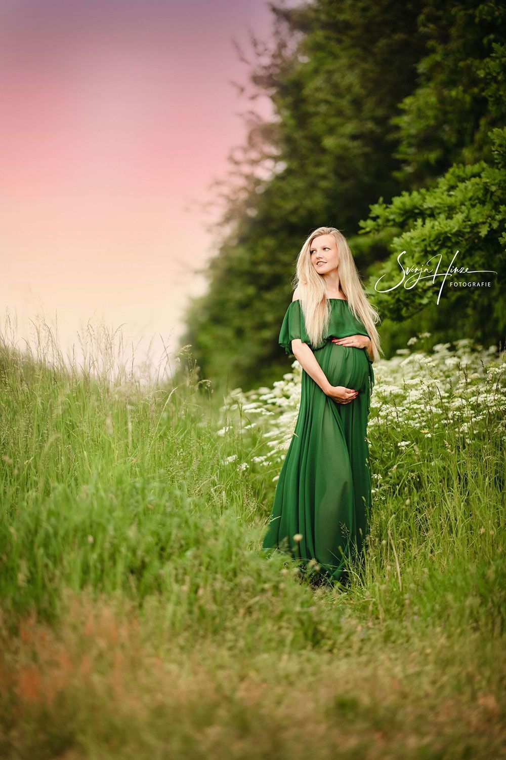 Eine schwangere Frau in einem grünen Kleid steht auf einem Feld mit hohem Gras.