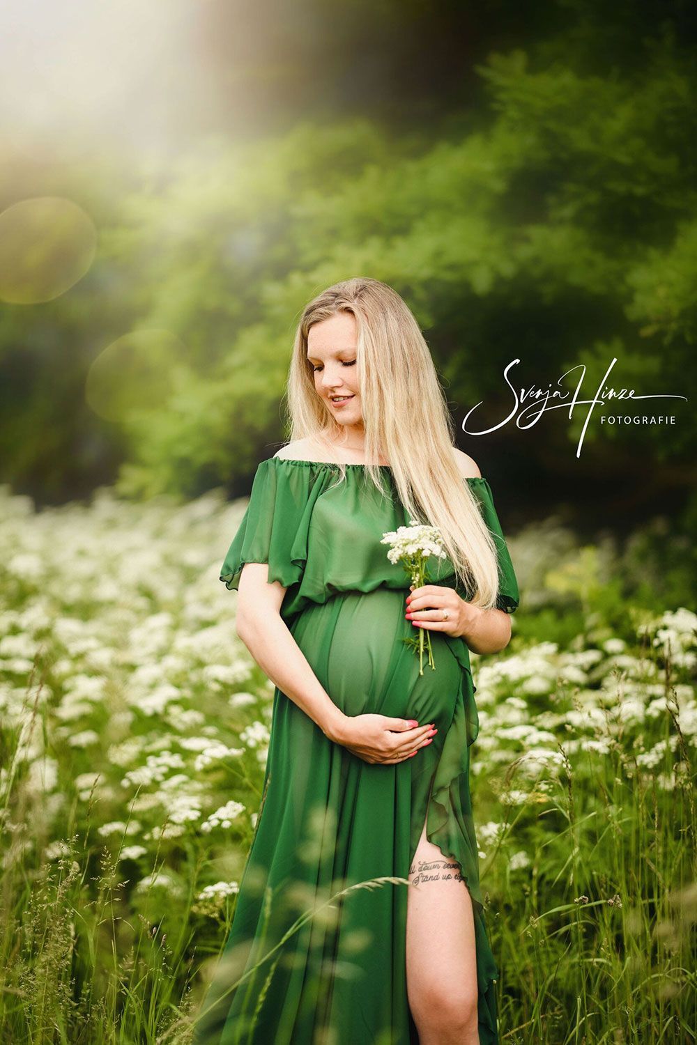 Eine schwangere Frau in einem grünen Kleid steht in einem Blumenfeld und hält eine Blume in der Hand.